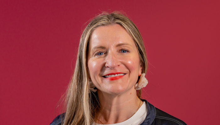 A woman with light brown hair smiles in front of a rubine background