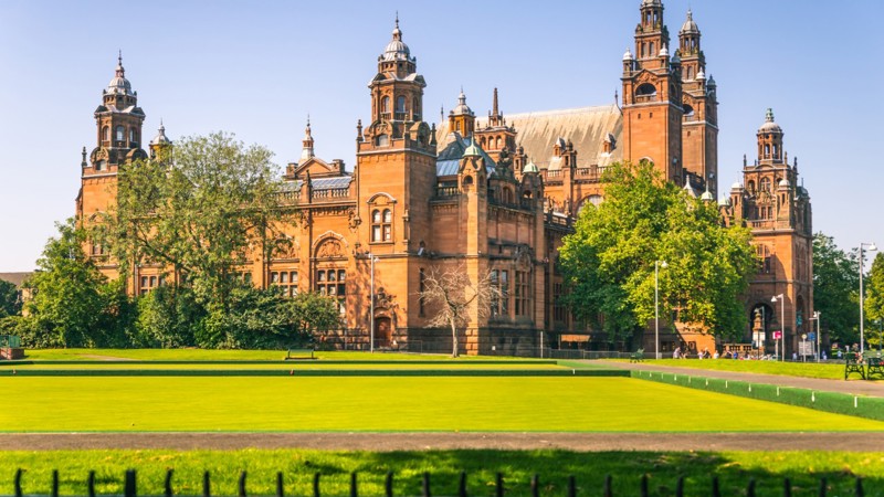 exterior shot of a red sandstone museum which is grand and has pillars at the front. the museum is surrounded by green trees and grass